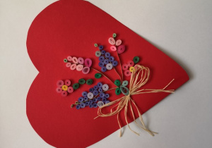 Zdjęcie przedstawia czerwone serce na białym tle. Na sercu znajduje się bukiet kolorowych kwiatów z żółtą kokardką.