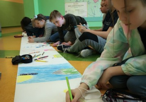 Uczniowie siedzą wzdłuż rozwiniętej rolki papieru i wykonują pracę plastyczną.