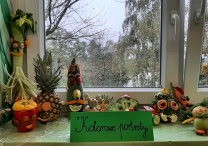 Postacie wykonane z owoców i warzyw ustawione na oknie. Przed pracami znajduje się kartka z napisem "Kolorowe portrety"