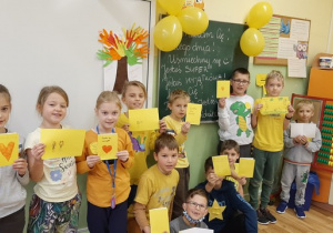 Uczniowie ustawieni w grupie. Trzymają żółte kartki – laurki. W tle sala lekcyjna oraz żółte balony.
