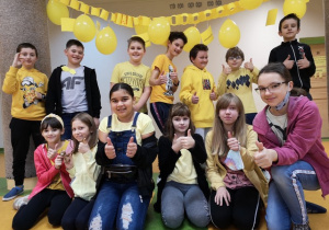 Uczniowie ustawieni w grupie. Uśmiechają się. Trzymają kciuki do góry. W tle szkolny korytarz oraz żółte balony.